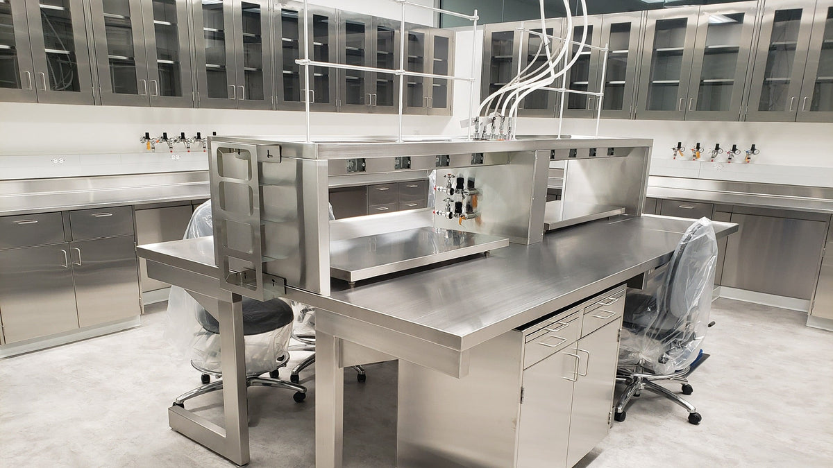 A large stainless steel laboratory work station with a long rectangular table, overhead utility frame, and two office-style chairs in a clean modern lab interior.