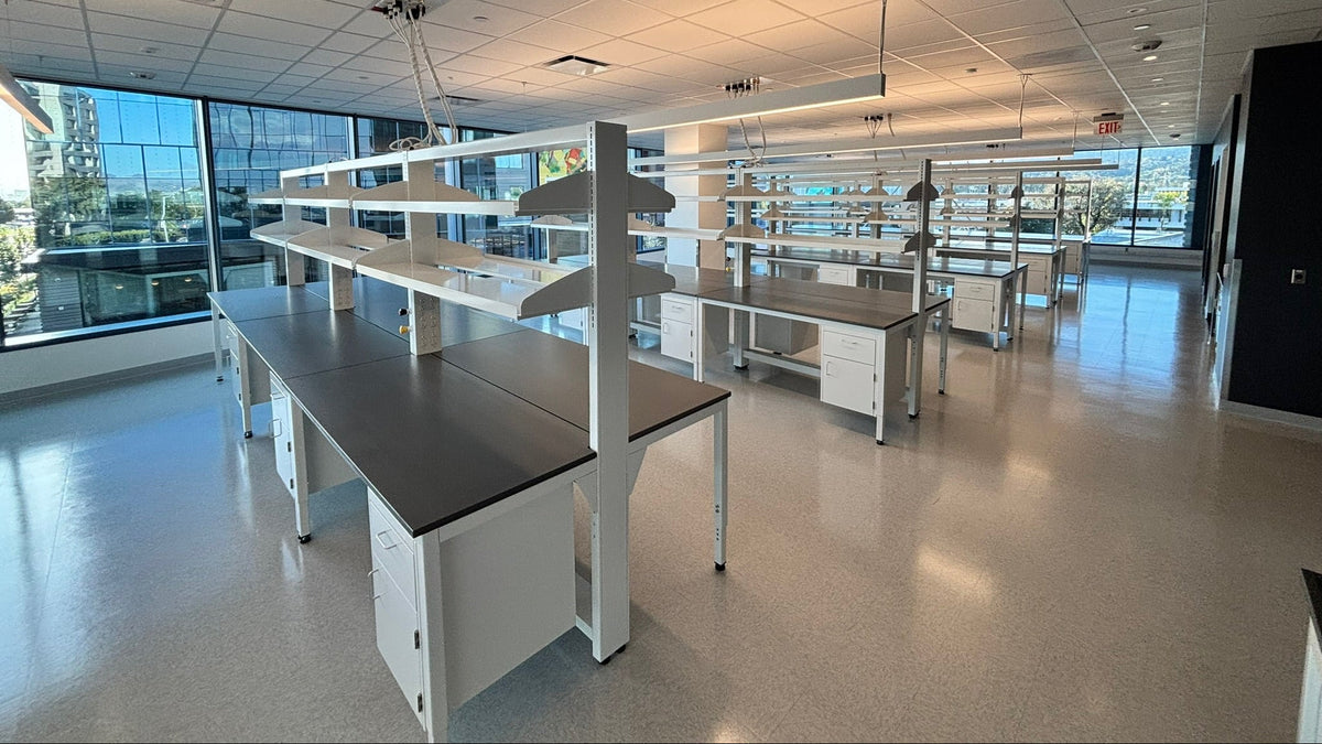 A large, modern laboratory or workshop space with multiple white workbenches, dark gray tops, shelving units, and tall windows on the left side showing an outdoor view.