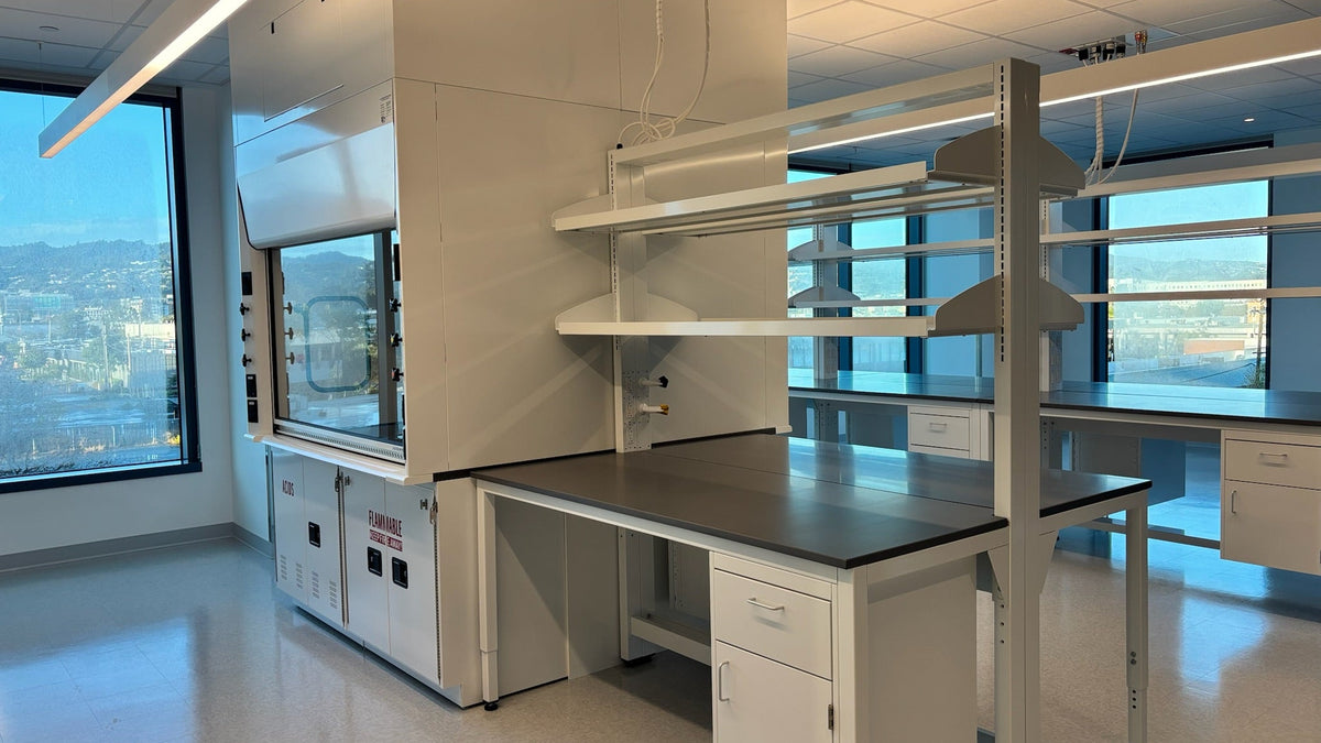 A modern white laboratory workstation setup with a dark gray workbench top, open shelving, and a biosafety hood, photographed in a bright, contemporary lab interior with large windows.